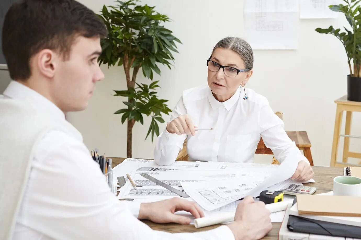 Seitliche Aufnahme eines nervösen jungen braunhaarigen männlichen Architekten, der am Schreibtisch mit Bauplänen sitzt und verängstigt ist, während eine wütende Chefin mittleren Alters ihn wegen Fehlern im Architekturplan ausschimpft.